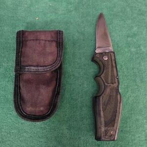 May include: A folding knife with a silver blade and a textured green and black handle, next to a dark brown sheath. The knife is open, revealing the sharp blade. The sheath is closed with a flap.