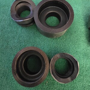 以下が含まれることがあります： さまざまな内径を持つ円形部品とねじ付き部品を含む、暗い金属製の工具のコレクション。工具は緑色のテクスチャ加工された表面に配置されており、作業場や機械的な環境を示唆しています。