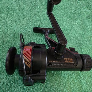 May include: A black fishing reel with the text "SILSTAR EX 40" and "SILSTAR" in red and gold. The reel has a handle and a spool. The background is a green surface.
