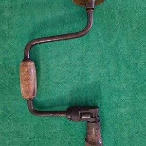 May include: A vintage hand brace tool featuring a dark brown metal crank frame, a round wooden top handle, a cylindrical wooden side handle, and a metal chuck mechanism at the base for holding drill bits, showing signs of age and wear.