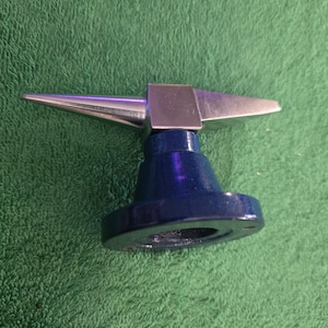 May include: A metal bench block anvil featuring a polished silver-colored top with a pointed horn on one side and a flat square surface on the other, mounted on a sturdy, circular, dark blue painted base with a hollow center for stability.