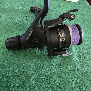 May include: Black FX4000 fishing reel with a purple and white spool. The reel has a handle and a gear ratio of 4.2:1. The reel has text that reads "LINE CAP. (Lbs.-Yds.) 8-240-10-200-12-160 SPOOL REMOVAL".