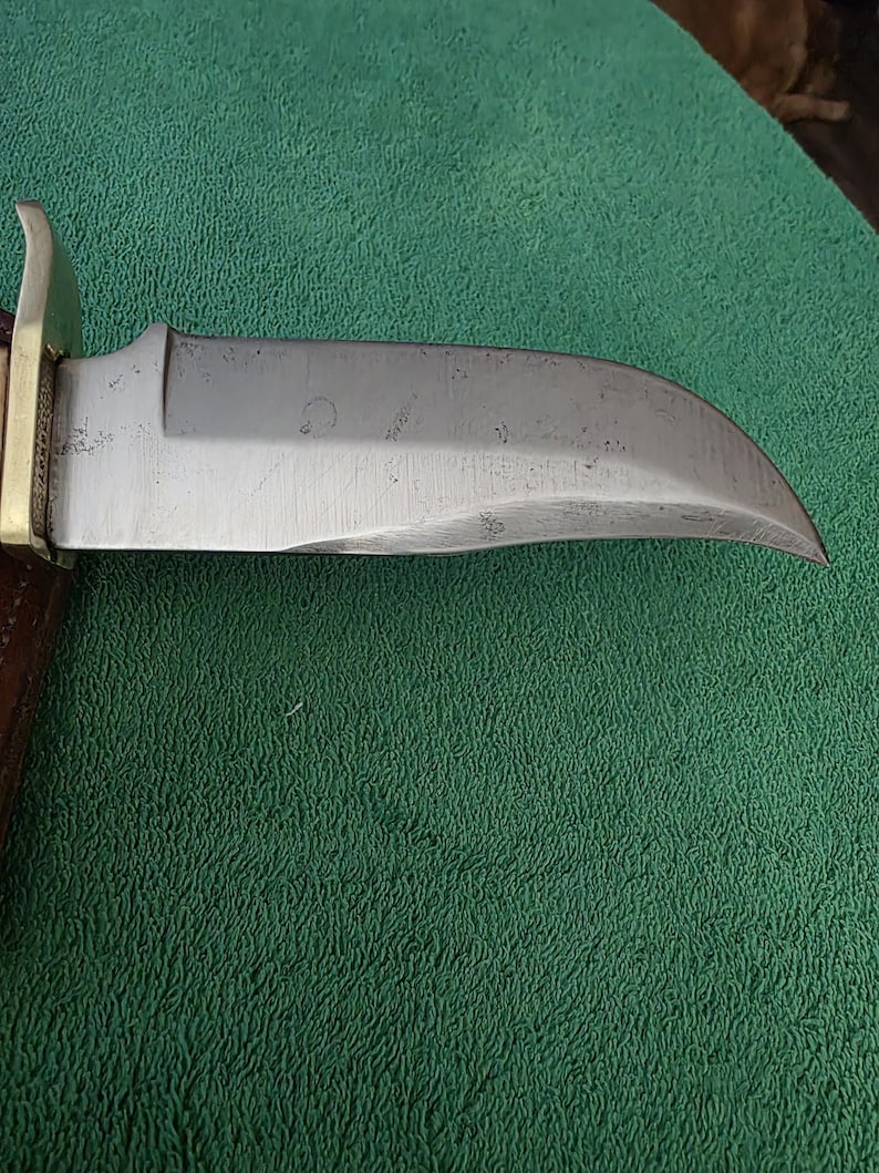 Pu&ograve; includere: Primo piano di una lama di coltello argentata con una leggera curva. La lama presenta alcuni piccoli graffi e una scritta appena visibile.