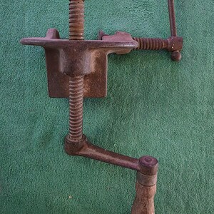 May include: An antique, rusty metal clamp with a wooden handle. The clamp features a screw mechanism and a flat base. The handle is made of wood and is attached to a lever. The clamp is designed to hold objects in place.