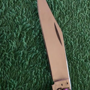 May include: Close-up of a stainless steel pocket knife with a partially open blade. The blade is sharp and reflects light, set against a green textured background. The knife's handle is visible, with a rounded end.