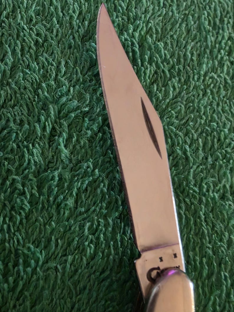 May include: Close-up of a stainless steel pocket knife with a partially open blade. The blade is sharp and reflects light, set against a green textured background. The knife's handle is visible, with a rounded end.