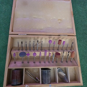 May include: A wooden box with an open lid, filled with various rotary tool bits and accessories. The bits are in different shapes and colors, including pink, brown, and silver. The box also contains two metal containers and a pair of tweezers.