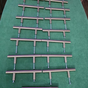May include: A collection of gold-toned T-bar cabinet pulls arranged on a green surface. Each pull has a cylindrical bar and two posts with screws for installation. The pulls are evenly spaced and ready for use.