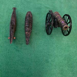 May include: A collection of three miniature metal figurines consisting of a submarine, a naval warship, and a field artillery cannon with two large black wheels. Each item features a dark metallic finish with visible structural details.
