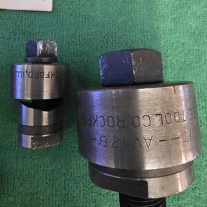 以下が含まれることがあります： 「ROCKFORD, ILL」と「UT--AV128--ROCKFORD TOOL CO」の文字が入った2つの金属製工具。工具は円筒形で、上部に六角ナット、下部にねじ山付きのベースが付いています。背景は緑色の布です。