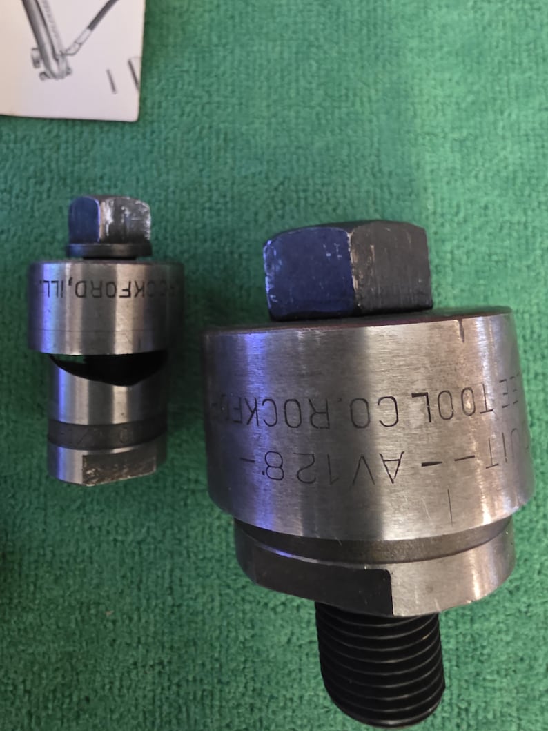 以下が含まれることがあります： 「ROCKFORD, ILL」と「UT--AV128--ROCKFORD TOOL CO」の文字が入った2つの金属製工具。工具は円筒形で、上部に六角ナット、下部にねじ山付きのベースが付いています。背景は緑色の布です。