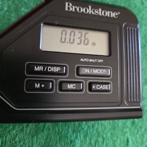 May include: A black Brookstone digital measuring device featuring an LCD screen displaying 0.036 m. The device includes buttons labeled MR/DISP, ON/MODE, M+, MC, and +CASE, with an AUTO SHUT OFF indicator printed above the ON/MODE button.