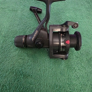 May include: A black fishing reel with the text "AX 2000" on the side. The reel has a handle, a spool, and various adjustment knobs. The reel is set against a green background.