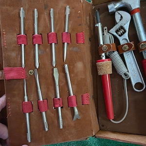 May include: A vintage brown leather tool kit with a variety of tools. The kit includes screwdrivers, a hammer, a wrench, and a soldering iron. The tools are secured with red fabric straps and leather holders. The hammer and soldering iron have red handles.
