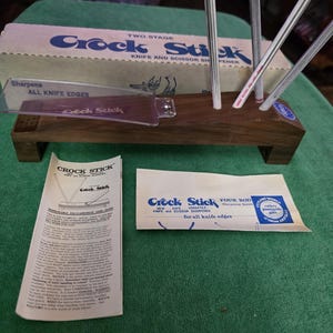 May include: A Crock Stick knife and scissor sharpener set. The set includes a wooden base with sharpening rods, a clear plastic guide, and printed instructions. The box and packaging feature the "Crock Stick" logo.