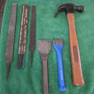 May include: A collection of hand tools including files, chisels, and a hammer, arranged on a green cloth. The tools are made of steel and wood, with some showing signs of use. One file is labeled "FULLER'S ALL-PURPOSE FILE".