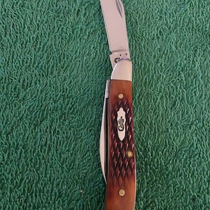 May include: A Case pocket knife with a partially open stainless steel blade. The handle is brown with a diamond pattern and brass rivets. The Case logo is visible on the handle. The knife is set against a green background.