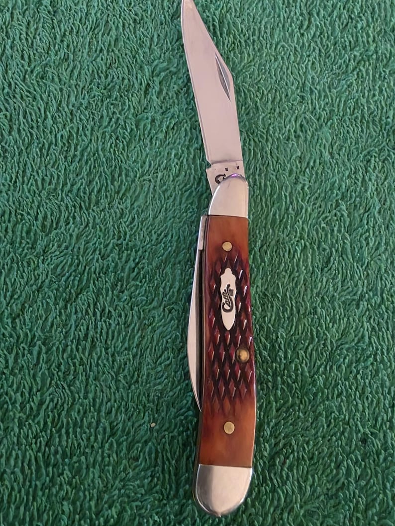May include: A Case pocket knife with a partially open stainless steel blade. The handle is brown with a diamond pattern and brass rivets. The Case logo is visible on the handle. The knife is set against a green background.