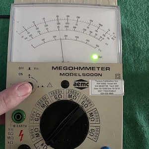 May include: A beige Megohmmeter, model 5000N, with a circular dial and a needle indicating a reading. The device has a black knob and various input terminals. The text "MEGOHMMETER" is visible, along with other technical markings.