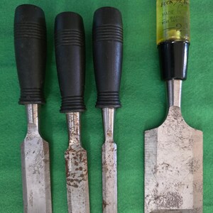 May include: A set of four metal wood chisels. Three chisels feature black ribbed handles, while the fourth chisel has a translucent yellow handle with the text BUCK BROS MADE IN USA WARNING WEAR SAFETY GLASSES 2 printed on the top section.