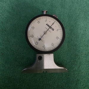 May include: Ames 11B-03PJ dial indicator with a black bezel and a white face. The dial reads from 0 to 25, with markings for increments of 0.0005 inches. The indicator is mounted on a silver-colored base.
