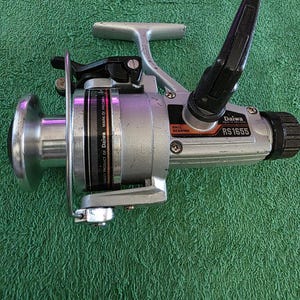 May include: Silver and black Daiwa RS1655 fishing reel. The reel has a silver handle, a black crank, and a black and orange label that reads "Ball Bearing". The reel is on a green surface.