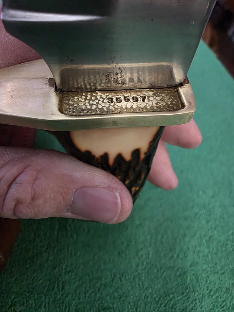 Pu&ograve; includere: Primo piano di un manico di coltello con una guardia in ottone e un manico in corno di cervo. La guardia ha il numero 35597 inciso.
