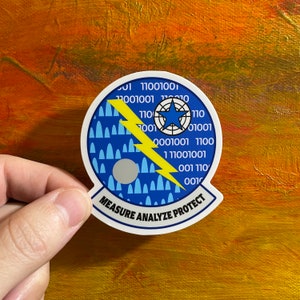 Op de afbeelding: Een witte sticker met een blauwe cirkel met daarin een gele bliksem, een blauwe ster en een cirkel met binaire code. De tekst "MEASURE ANALYZE PROTECT" staat rond de cirkel.