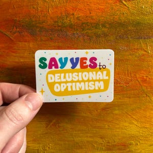 Può includere: Sticker bianco con testo colorato che recita "SAY YES to DELUSIONAL OPTIMISM". Lo sticker ha uno sfondo giallo con puntini bianchi e stelle.