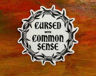 Può includere: Un adesivo bianco con la scritta "Cursed with Common Sense" in nero, con un carattere gotico. Il testo è circondato da un disegno di filo spinato. L'adesivo è su uno sfondo dai toni arancioni e marroni.