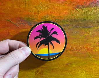 Puede incluir: Una pegatina redonda con una escena de puesta de sol con una silueta negra de palmera. El cielo transiciona de rosa a naranja, con un sol amarillo que se pone sobre un océano azul. La pegatina tiene un borde negro.