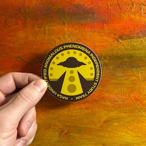 May include: A yellow and brown round sticker with the text "NASA Unidentified Anomalous Phenomena Independent Study Team" and a stylized image of a UFO with a beam of light.