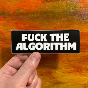 May include: A rectangular black sticker featuring white sans-serif text which reads FUCK THE ALGORITHM in two lines. The sticker has a clean white border around the edges and is held against a textured orange and yellow painted background.