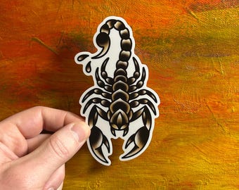 May include: A sticker with a scorpion design, featuring a gradient of black and brown. The scorpion has a curved tail and large pincers. The sticker is held against an orange and yellow background.