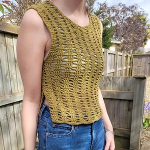 May include: A sleeveless crochet top in a mustard yellow color. The top has a loose, open weave and a slightly cropped length. The top is worn over a pair of blue jeans.