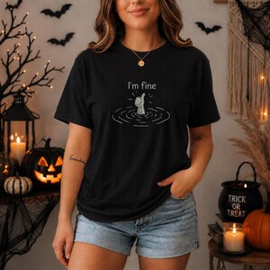 May include: A black short-sleeve t-shirt featuring a white graphic of a hand emerging from water giving a thumbs-up gesture. Above the hand, the text reads I'm fine in a simple white font, surrounded by circular ripples in the water.
