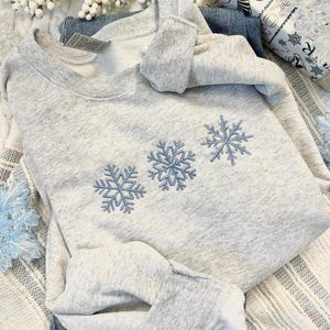 May include: A light gray sweatshirt with three embroidered blue snowflakes. The sweatshirt is folded on top of a pair of blue jeans. The image is styled with winter-themed decorations.
