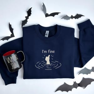 May include: A navy blue long-sleeve sweatshirt featuring an embroidered graphic of a hand giving a thumbs-up from water with the text I'm fine. Beside it sits a black mug with red interior and the white text Vampire Juice, surrounded by paper bats.