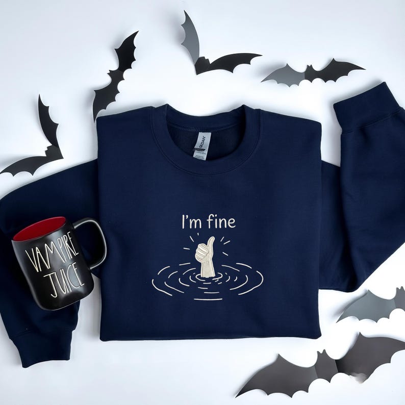 May include: A navy blue long-sleeve sweatshirt featuring an embroidered graphic of a hand giving a thumbs-up from water with the text I'm fine. Beside it sits a black mug with red interior and the white text Vampire Juice, surrounded by paper bats.