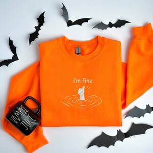 May include: A bright orange long-sleeve sweatshirt featuring an embroidered white graphic of a hand giving a thumbs-up gesture emerging from water ripples with the text I'm fine above it, accompanied by a black mug labeled VAMPIRE JUICE.