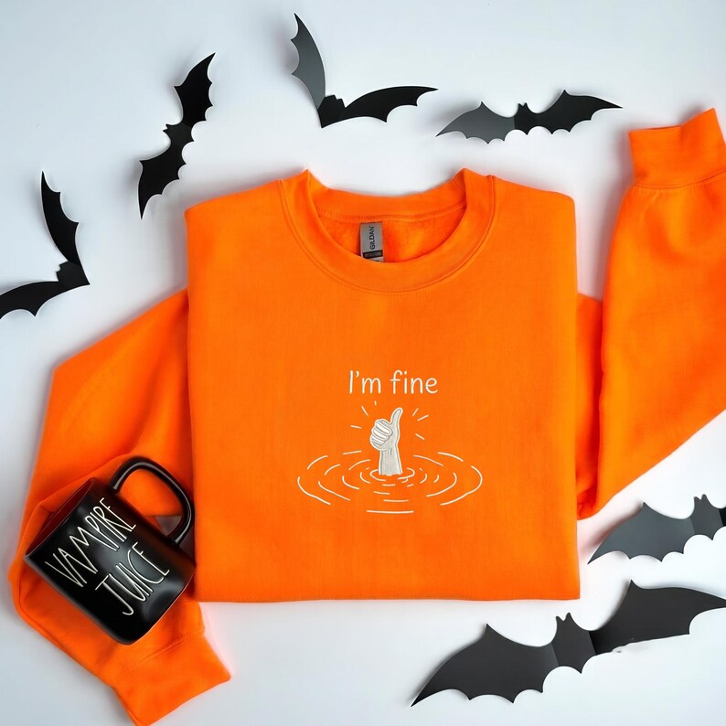 May include: A bright orange long-sleeve sweatshirt featuring an embroidered white graphic of a hand giving a thumbs-up gesture emerging from water ripples with the text I'm fine above it, accompanied by a black mug labeled VAMPIRE JUICE.
