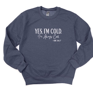 May include: A navy blue sweatshirt with the text "YES, I'M COLD. I'm Always Cold. -ME 24/7" in white. The sweatshirt has a crew neck and long sleeves. A casual, comfortable garment for everyday wear.