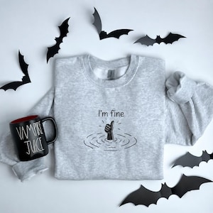 May include: A light gray crewneck sweatshirt featuring a black embroidered graphic of a hand giving a thumbs up from water with the text I'm fine above it, accompanied by a black mug with the white text Vampire Juice and black paper bats.