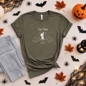 May include: An olive green short-sleeved t-shirt featuring a white graphic of a hand emerging from water giving a thumbs-up gesture. Above the hand, the text reads I'm fine in a simple white font. The shirt has a crew neckline and short sleeves.