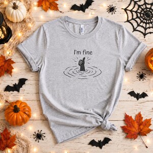 May include: A light gray short-sleeve t-shirt featuring a black graphic of a hand emerging from water giving a thumbs-up gesture, accompanied by the text "I'm fine" printed in a simple black font above the illustration on the center chest.