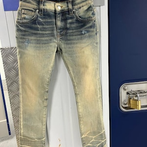 Puede incluir: Vaqueros de mezclilla azul claro desgastados con un lavado amarillo desteñido. Los vaqueros tienen un ajuste delgado y están colgados de una percha de madera. La etiqueta de la marca "AMIRI" es visible en la cintura.