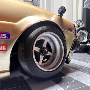 May include: A gold-colored model car with a black tire and silver rim. The car has a "WAKOS" and "MOTUL" sticker on the side. The car is sitting on a black and white checkered floor.