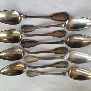Könnte beinhalten: Ein Set aus acht silberfarbenen Metalllöffeln, die in vier Paaren angeordnet sind. Jeder Löffel hat eine längliche, spitz zulaufende Laffe und einen Griff mit einem dekorativen, abgerundeten Ende. Die Metalloberflächen weisen natürliche Patina auf.