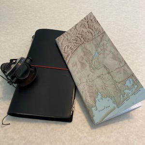 May include: A black leather notebook with a red elastic band and a zipper closure. A map of a coastal area is folded and placed on top of the notebook.