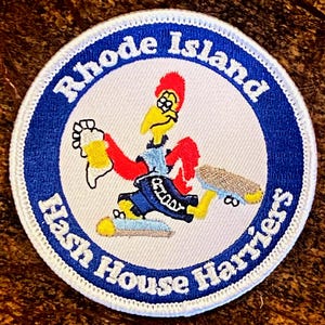 May include: A white, round patch with a blue circle in the center. The blue circle has white text that reads "Rhode Island Hash House Harriers". Inside the blue circle is a cartoon rooster wearing a blue and red outfit, holding a beer bottle and a skateboard.